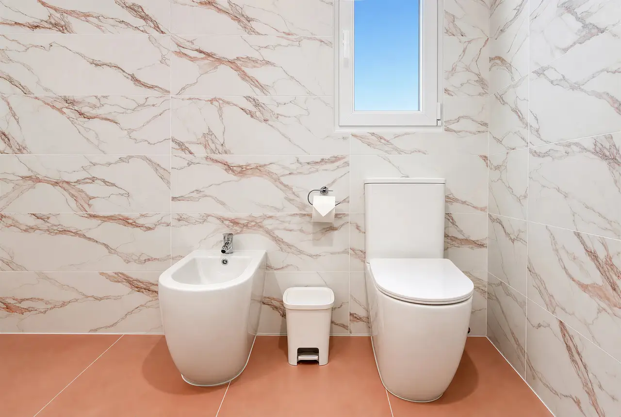 Toilet, bidet and small window in a bathroom with pink-veined tiles