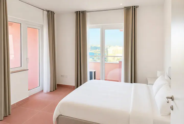 Double bed with white linen, a window and glass doors onto a pink balcony